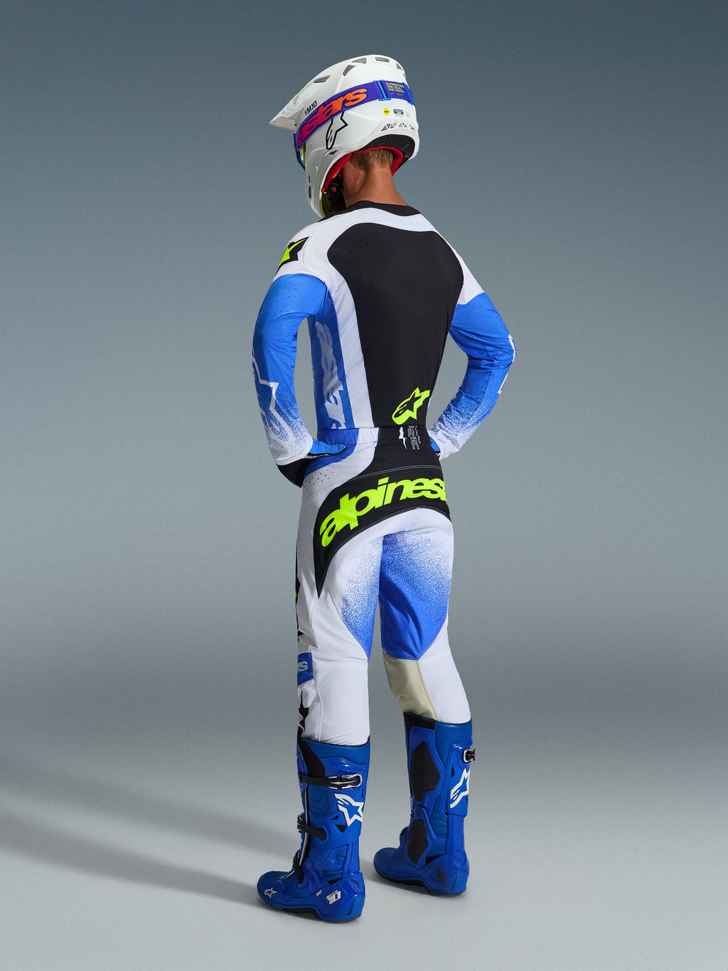 Piloto de motocross visto de costas com o conjunto de equipamento completo Alpinestars 2026 Techstar Knif em Azul Ucla e preto, incluindo uma camisola com painel traseiro preto e logótipo amarelo néon, calças azuis e brancas com branding amarelo vibrante, botas de motocross azuis e capacete de corrida branco.