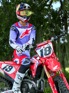 Piloto de motocross numa moto Honda CRF vermelha, a usar a camisola Alpinestars 2025 Fluid Wurx em roxo e vermelho brilhante, calças a condizer em roxo e vermelho, capacete Red Bull com óculos amarelos, botas e luvas Alpinestars brancas, equipamento profissional de MX para condução todo-o-terreno