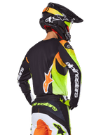 Piloto de motocross visto de costas com o conjunto de equipamento Alpinestars 2025 Fluid Wurx, camisola preta com gráficos vibrantes em amarelo néon e laranja, calças a condizer com painéis brancos e laranja, capacete preto com detalhes néon, logótipo Alpinestars em destaque nas mangas e no assento