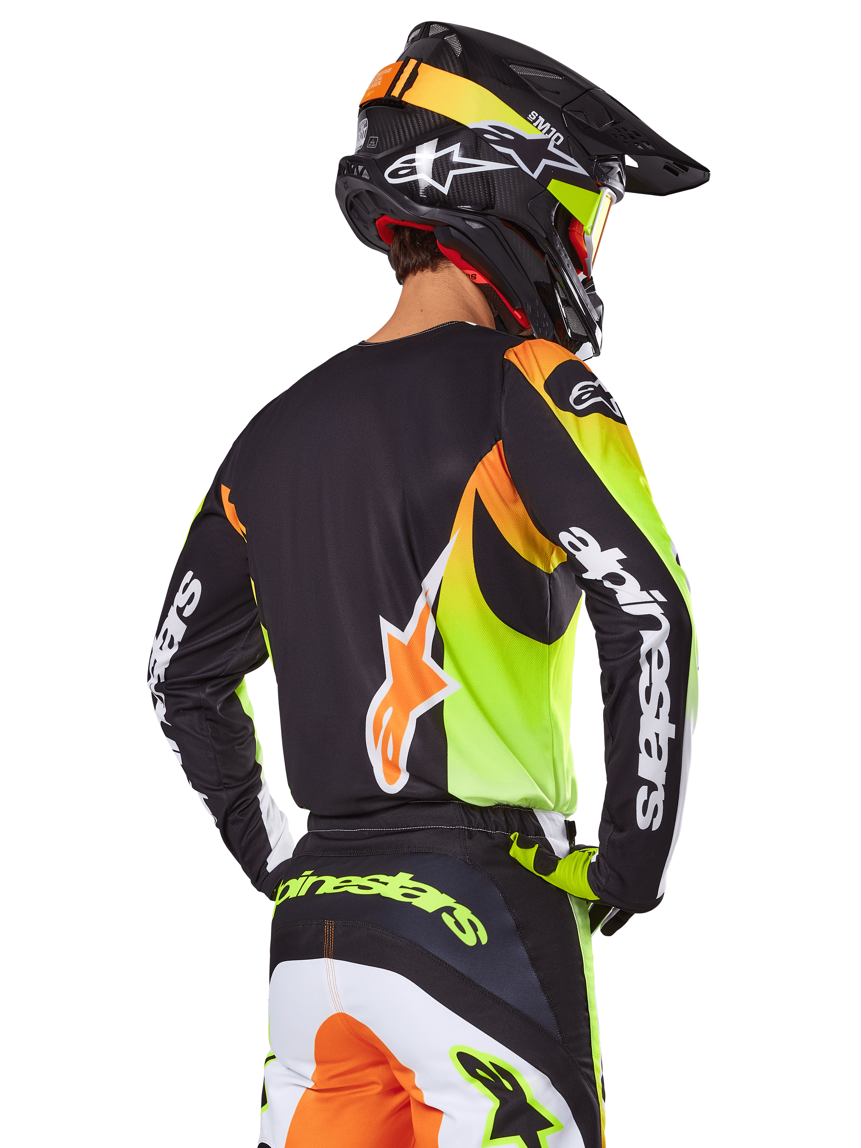 Piloto de motocross visto de costas com o conjunto de equipamento Alpinestars 2025 Fluid Wurx, camisola preta com gráficos vibrantes em amarelo néon e laranja, calças a condizer com painéis brancos e laranja, capacete preto com detalhes néon, logótipo Alpinestars em destaque nas mangas e no assento