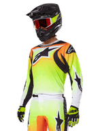Piloto de motocross a usar o Conjunto de Equipamento Alpinestars 2025 Fluid Wurx, camisola com gradiente amarelo e laranja, detalhes pretos e logótipos de estrela grandes, calças de motocross a condizer, capacete integral preto com óculos de lentes vermelhas e luvas amarelas, conjunto de alta performance para todo-o-terreno