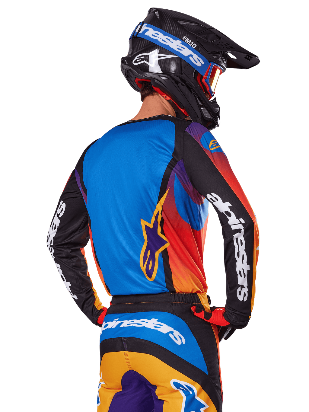 Piloto de motocross visto de costas a usar o conjunto de equipamento Alpinestars 2025 Fluid Wurx, camisola multicolor com costas azuis e mangas pretas com logótipos brancos, calças a condizer em laranja e azul, capacete preto e azul com óculos laranja, fato de corrida todo-o-terreno coordenado.