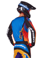 Piloto de motocross visto de costas a usar o conjunto de equipamento Alpinestars 2025 Fluid Wurx, camisola multicolor com costas azuis e mangas pretas com logótipos brancos, calças a condizer em laranja e azul, capacete preto e azul com óculos laranja, fato de corrida todo-o-terreno coordenado.
