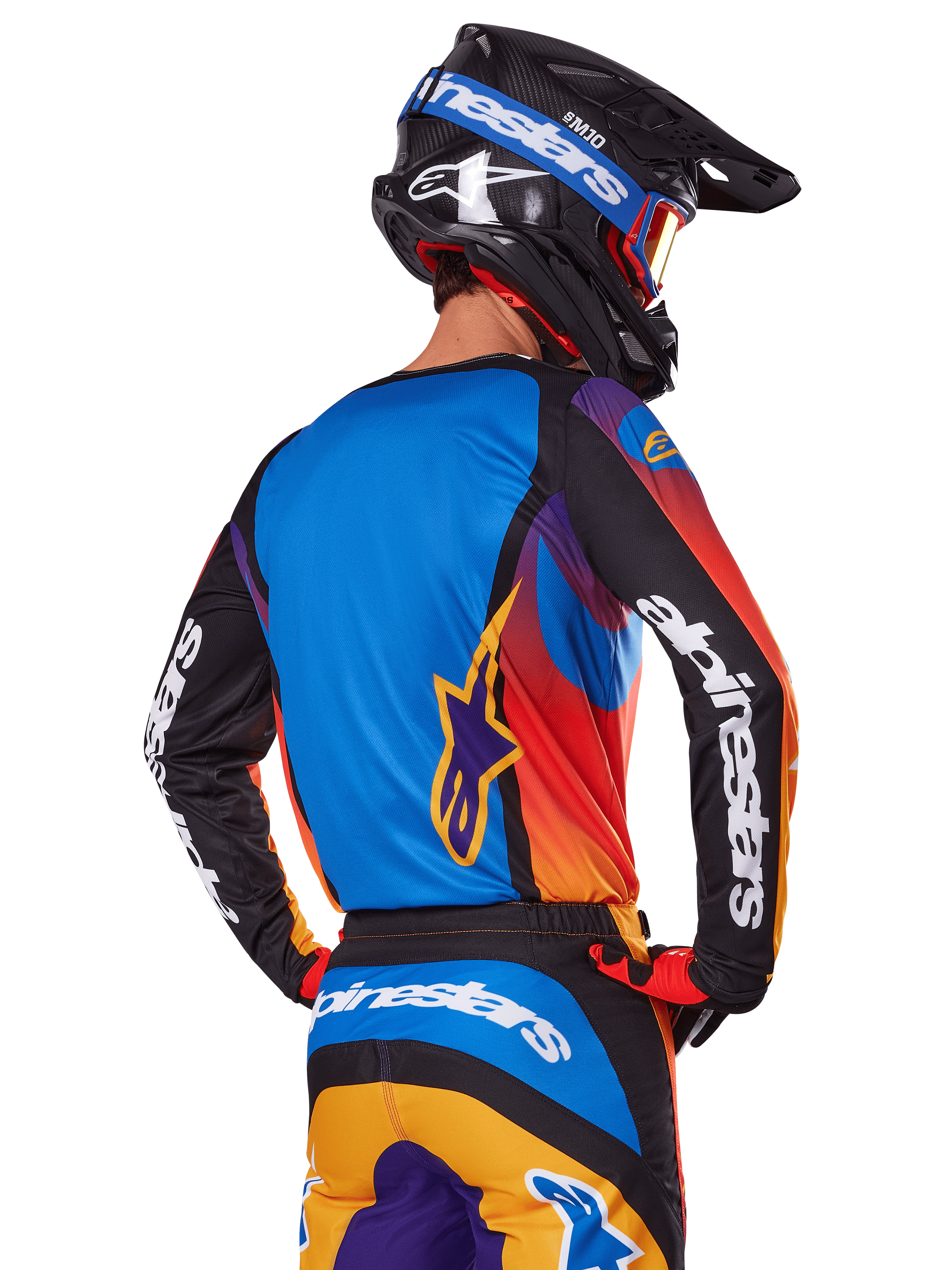 Piloto de motocross visto de costas a usar o conjunto de equipamento Alpinestars 2025 Fluid Wurx, camisola multicolor com costas azuis e mangas pretas com logótipos brancos, calças a condizer em laranja e azul, capacete preto e azul com óculos laranja, fato de corrida todo-o-terreno coordenado.