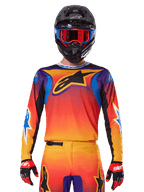 Alpinestars 2025 Fluid Wurx Jersey, camisola de motocross multicolor com gradiente laranja e vermelho e pormenores roxos, usada com equipamento a condizer, capacete preto com óculos azuis e vermelhos e luvas vermelhas, apresentando um grande logótipo de estrela preto no peito.