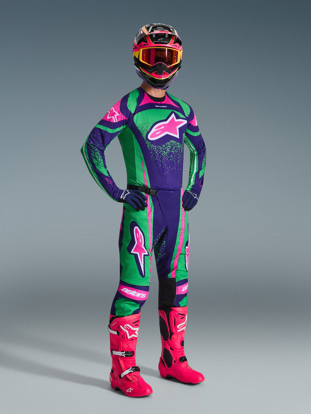 Piloto de motocross com o conjunto Alpinestars 2026 Techstar Nomur em roxo escuro, verde e rosa fluo, com camisola e calças a condizer, capacete colorido com óculos cor-de-rosa e botas cor-de-rosa vibrantes, equipamento profissional de competição todo-o-terreno