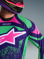 Primeiro plano da camisola Alpinestars Techstar Nomur 2026, em roxo profundo com detalhes em verde e rosa fluo, apresentando um grande logótipo de estrela cor-de-rosa no peito e perfurações cortadas a laser no ombro, vestida por um piloto com um capacete de motocross em roxo e rosa a condizer.
