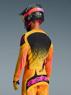 Camisola Alpinestars 2026 Techstar Nomur em Laranja, Amarelo e Rosa, vista traseira de um piloto de motocross com equipamento completo, incluindo calças a condizer e um capacete preto, design vibrante com detalhes em amarelo e rosa e painel traseiro preto, corte atlético de competição para performance em off-road