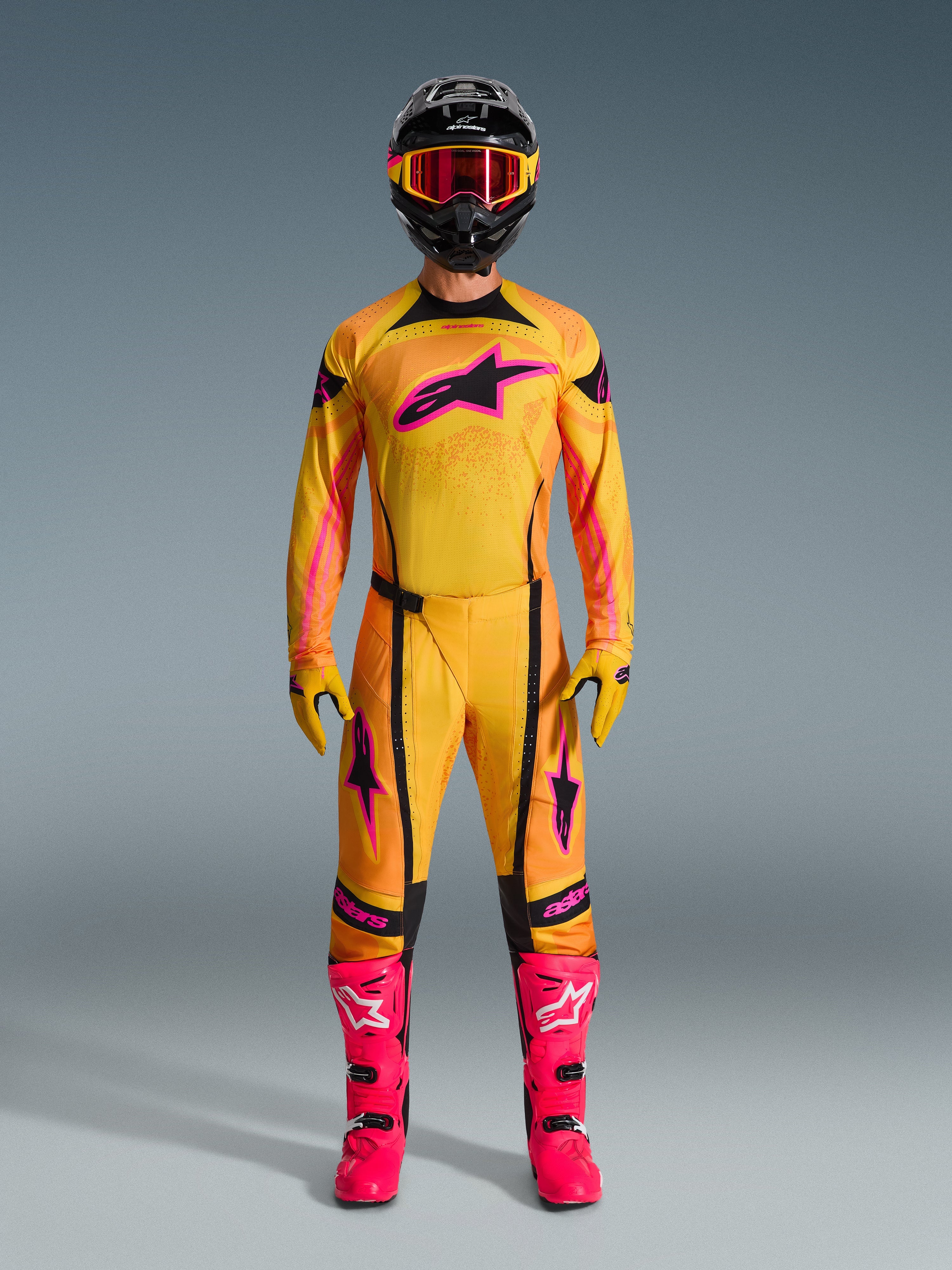 Piloto de motocross com a Camisola Techstar Nomur 2026 em laranja, amarelo e rosa, coordenada com calças de motocross a condizer, capacete preto com óculos rosa e botas rosa vibrante, equipamento com corte atlético de corrida para competição off-road