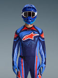 Piloto de motocross a usar a camisola Alpinestars 2026 Techstar Nomur em azul-marinho, laranja e azul UCLA, calças a condizer e luvas azuis, capacete azul com óculos brancos, logótipo Alpinestars grande em vermelho e branco no peito, equipamento de performance coordenado para corridas off-road.