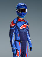 Piloto de motocross a usar a Camisola Alpinestars 2026 Techstar Nomur, equipamento de corrida em azul marinho, laranja e azul UCLA, com um grande logótipo Astars laranja, capacete e luvas azuis a condizer, conjunto de competição off-road atlético