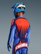 Piloto de motocross visto de costas com a Camisola Alpinestars Techstar Nomur 2026, em azul marinho com detalhes laranja e UCLA Blue, apresentando um grande grafismo texturado laranja nas costas, calças de motocross a condizer e capacete de corrida azul, equipamento profissional coordenado para condução off-road.