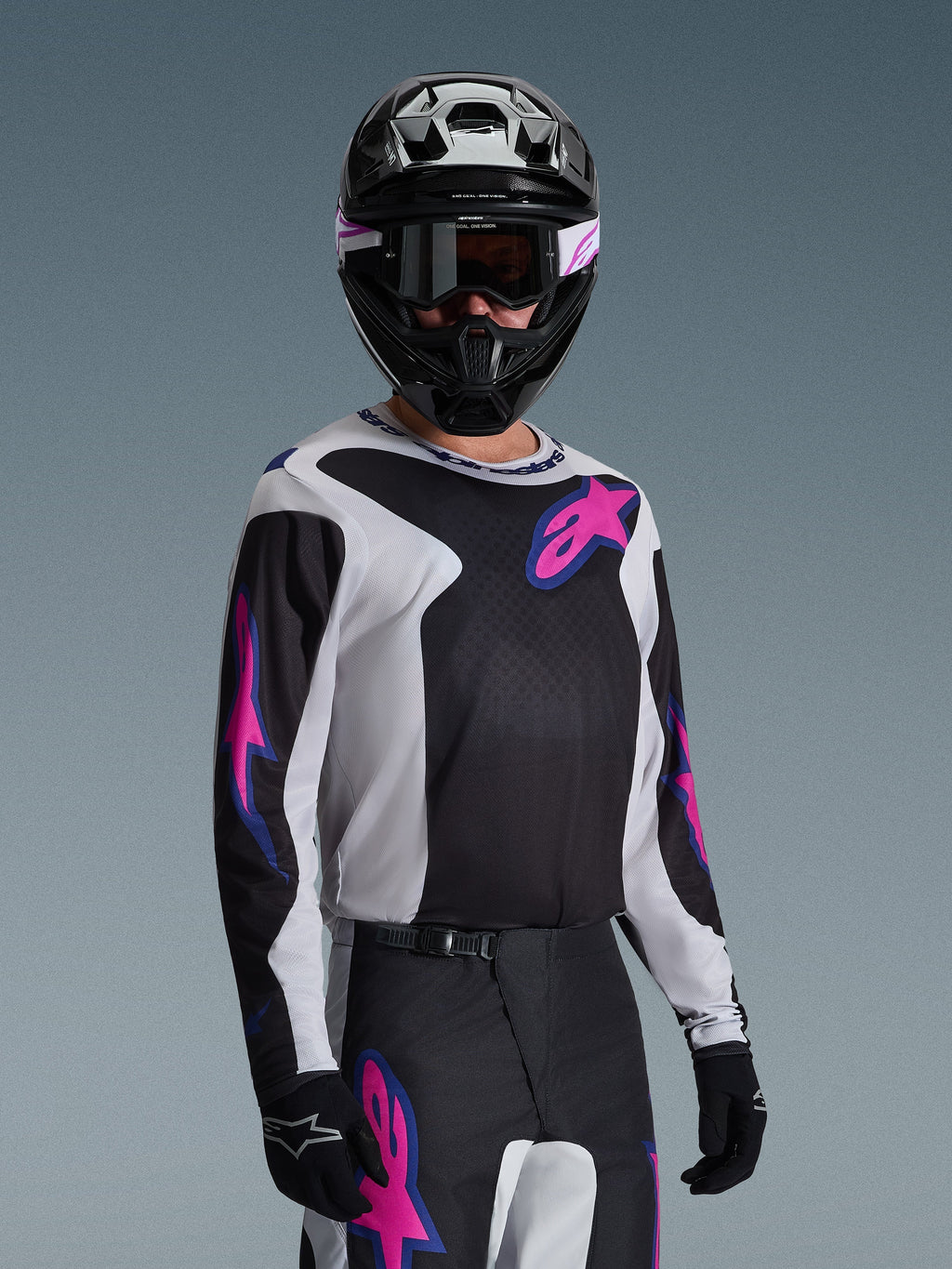 Piloto de motocross com camisola Alpinestars 2026 Fluid Grid em preto, cinzento claro e roxo, com grandes logótipos de estrela rosa e roxo, calças a condizer, capacete preto e óculos de proteção, conjunto completo de equipamento de corrida todo-o-terreno