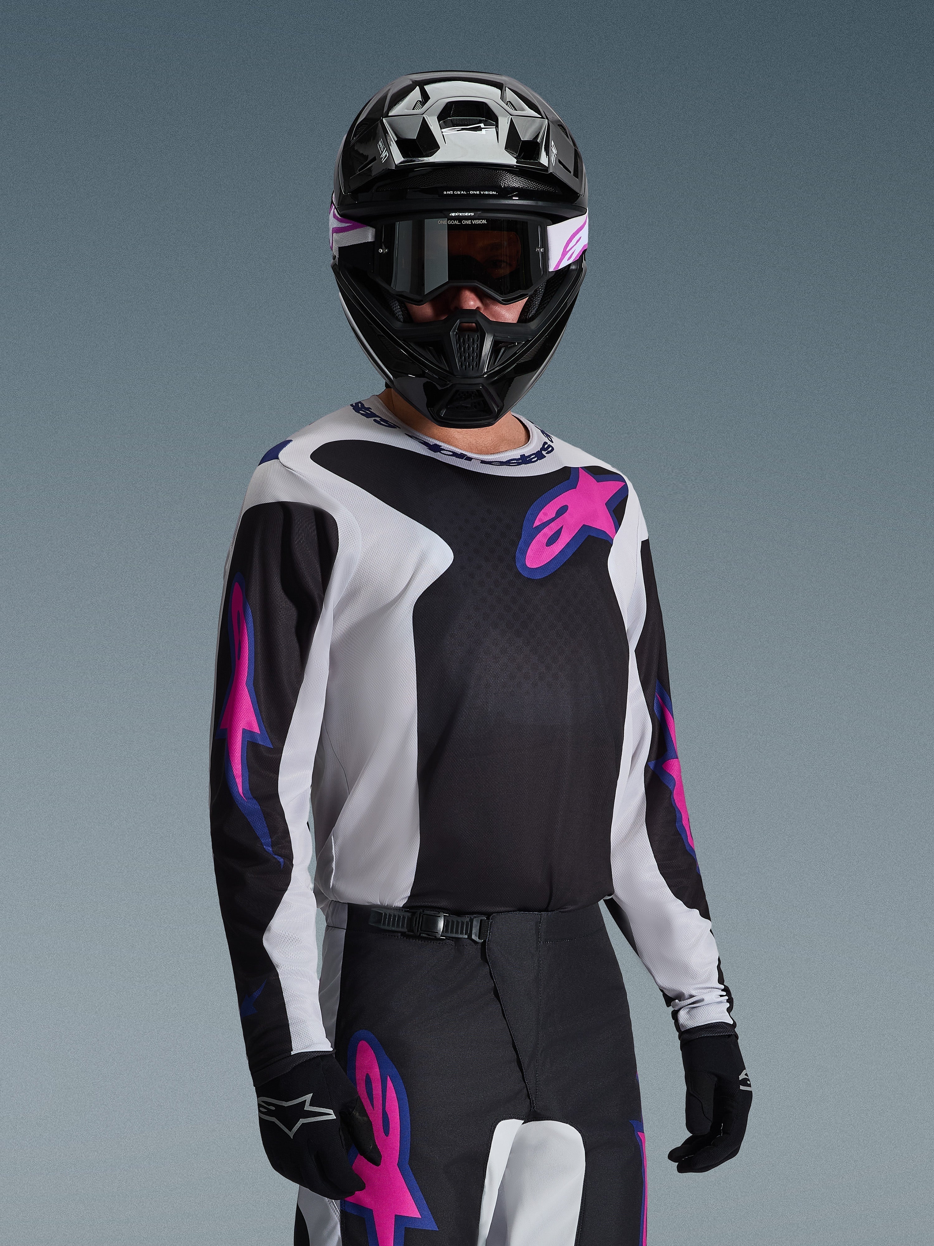 Piloto de motocross com camisola Alpinestars 2026 Fluid Grid em preto, cinzento claro e roxo, com grandes logótipos de estrela rosa e roxo, calças a condizer, capacete preto e óculos de proteção, conjunto completo de equipamento de corrida todo-o-terreno