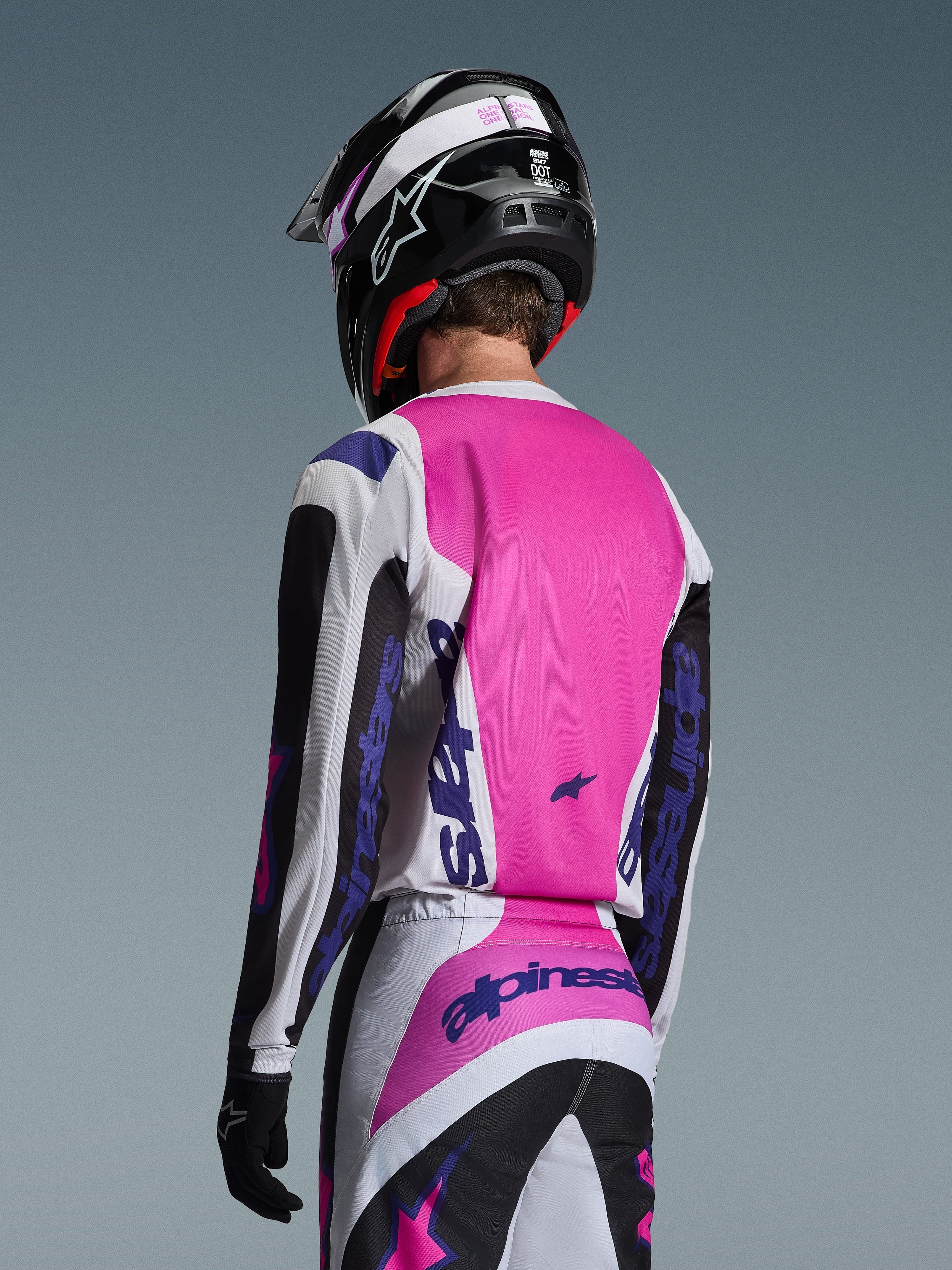 Piloto de motocross visto de costas a usar a camisola Alpinestars 2026 Fluid Grid em preto, branco e roxo, calças a condizer com logótipos destacados, capacete preto com detalhes cor-de-rosa e luvas pretas, equipamento completo coordenado para desempenho todo-o-terreno