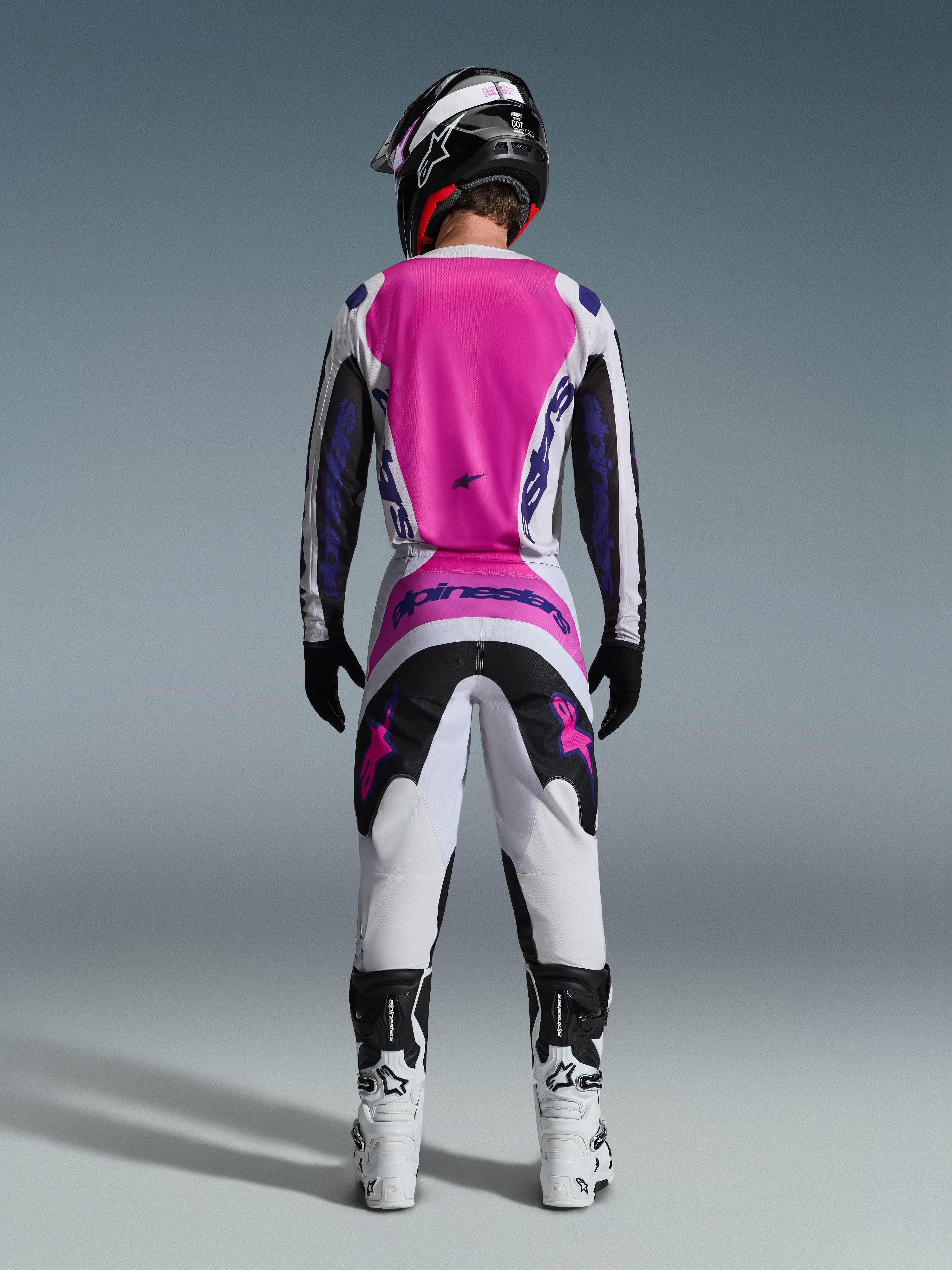 Alpinestars 2026 Fluid Grid Jersey e calças a condizer, conjunto de equipamento de motocross, rosa, branco e roxo, vista traseira mostrando o ajuste atlético ergonómico, usado com um capacete preto e botas brancas, concebido para desempenho todo-o-terreno.