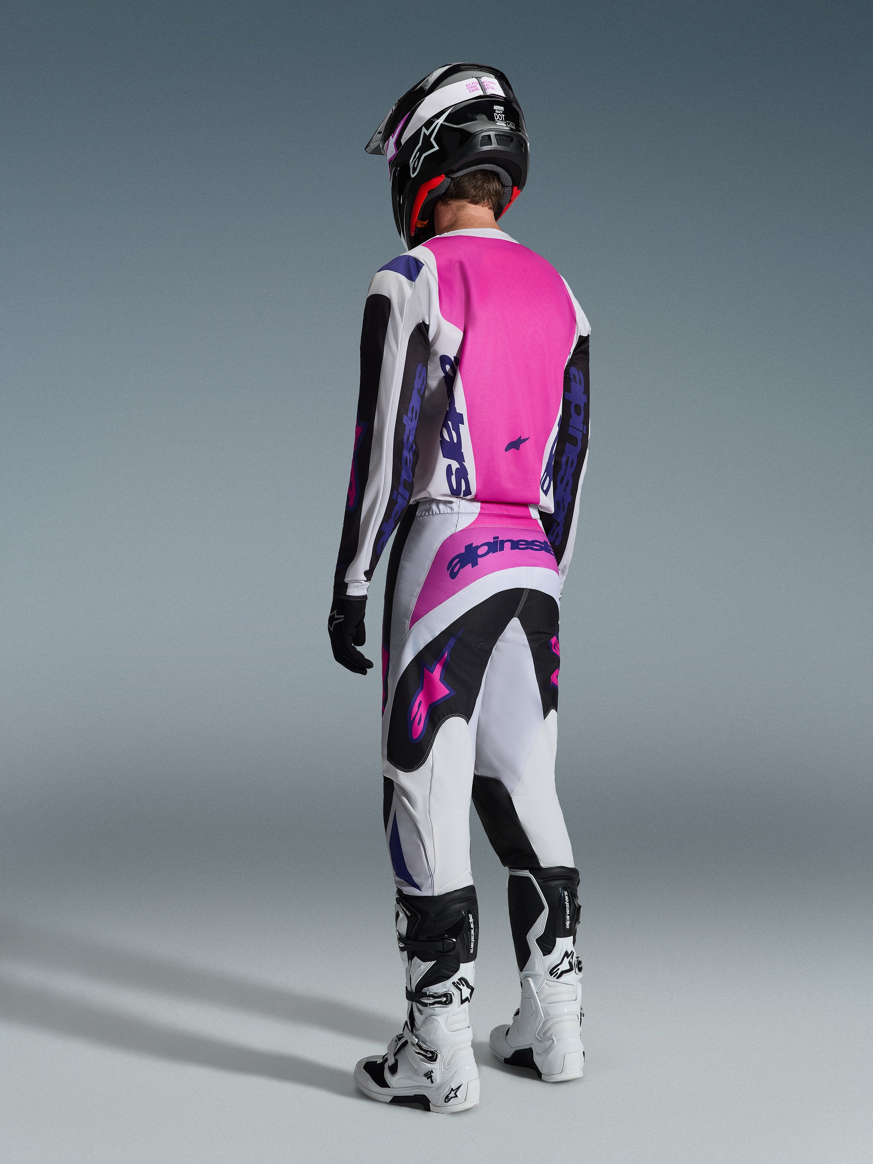 Piloto de motocross visto de costas a usar a camisola Alpinestars Fluid Grid 2026 e calças a condizer, em cinzento claro e preto com um painel traseiro rosa vibrante e logótipos roxos, completo com capacete preto e botas brancas