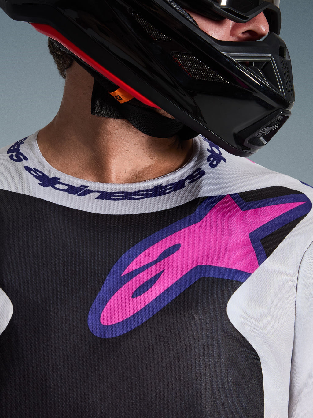 Grande plano da Camisola Alpinestars 2026 Fluid Grid, camisola de motocross, em preto, cinzento claro e roxo, com um logótipo de estrela vibrante em rosa e roxo no peito e gola desportiva com a marca, usada com um capacete de motocross preto