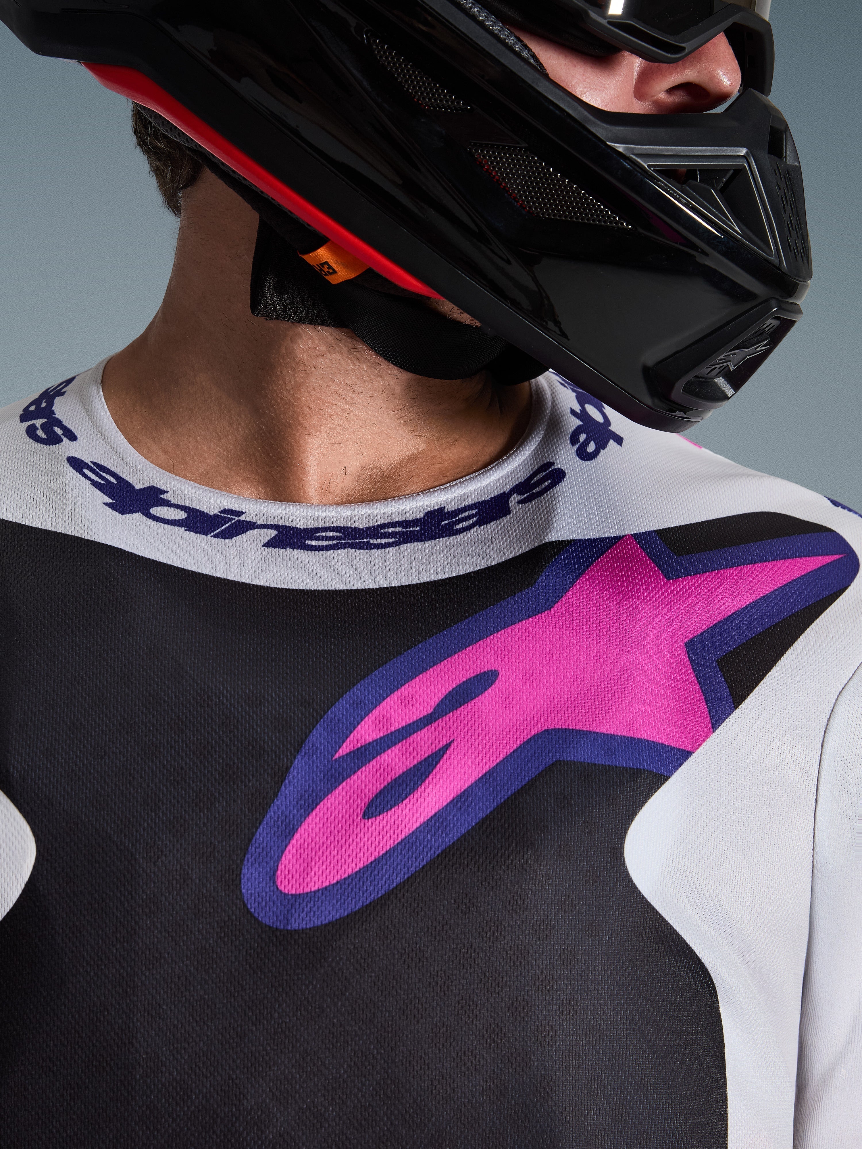 Grande plano da Camisola Alpinestars 2026 Fluid Grid, camisola de motocross, em preto, cinzento claro e roxo, com um logótipo de estrela vibrante em rosa e roxo no peito e gola desportiva com a marca, usada com um capacete de motocross preto
