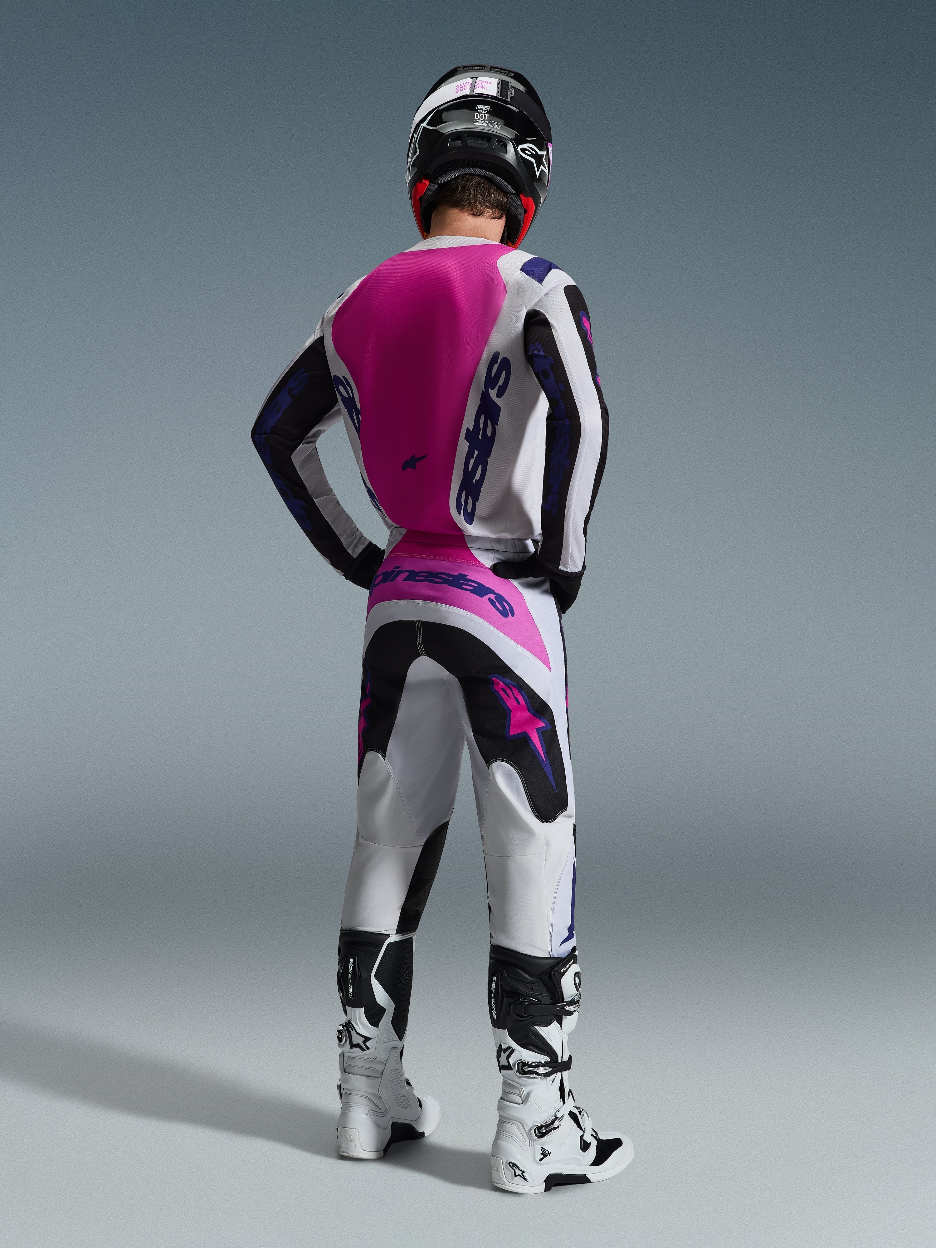 Piloto de motocross visto de costas a usar a camisola Alpinestars Fluid Grid 2026, camisola branca com um grande painel traseiro cor-de-rosa choque e logótipos roxos, calças brancas e pretas a condizer com detalhes cor-de-rosa, botas brancas e capacete preto, equipamento de corrida todo-o-terreno coordenado