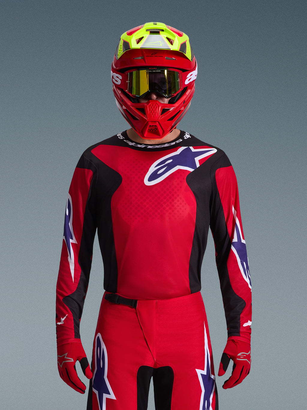Piloto de motocross com o conjunto de equipamento Alpinestars 2026 Fluid Grid, camisola vermelha com painéis laterais pretos e logótipo de estrela branco, calças e luvas vermelhas a condizer, capacete vermelho e amarelo com óculos de lentes douradas, equipamento de alta performance para todo-o-terreno