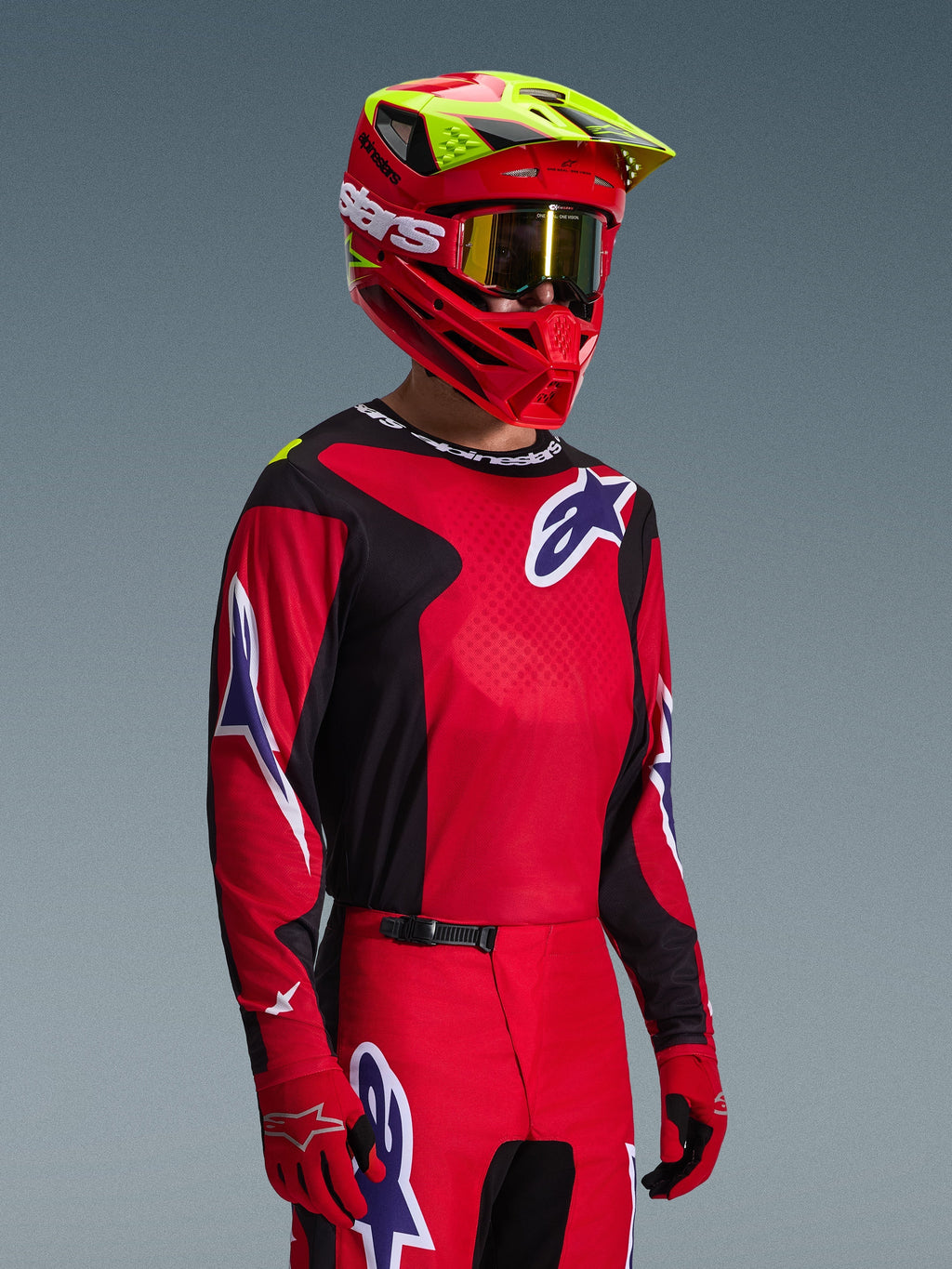 Piloto de motocross a usar o conjunto de equipamento Alpinestars Fluid Grid 2026, camisola vermelha e preta com detalhes do logótipo em branco e azul, calças vermelhas e pretas a condizer, capacete vermelho com realces amarelos e óculos dourados, equipamento de corrida todo-o-terreno coordenado
