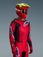 Piloto de motocross com a camisola Alpinestars Fluid Grid 2026 em vermelho, preto e roxo, calças de motocross coordenadas, capacete vermelho com pormenores em amarelo néon, óculos dourados e luvas vermelhas, equipamento completo de competição todo-o-terreno.