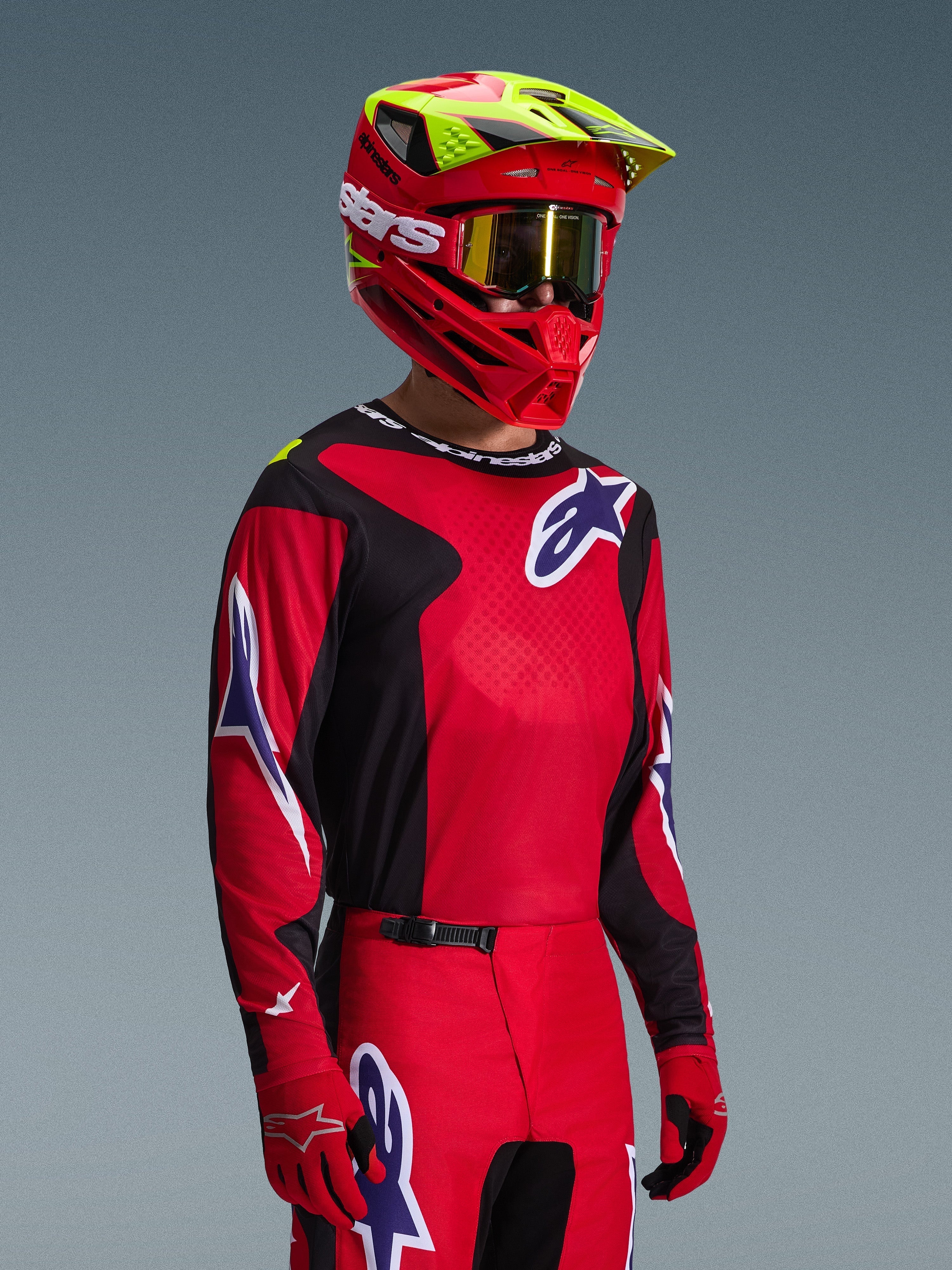 Piloto de motocross com a camisola Alpinestars Fluid Grid 2026 em vermelho, preto e roxo, calças de motocross coordenadas, capacete vermelho com pormenores em amarelo néon, óculos dourados e luvas vermelhas, equipamento completo de competição todo-o-terreno.