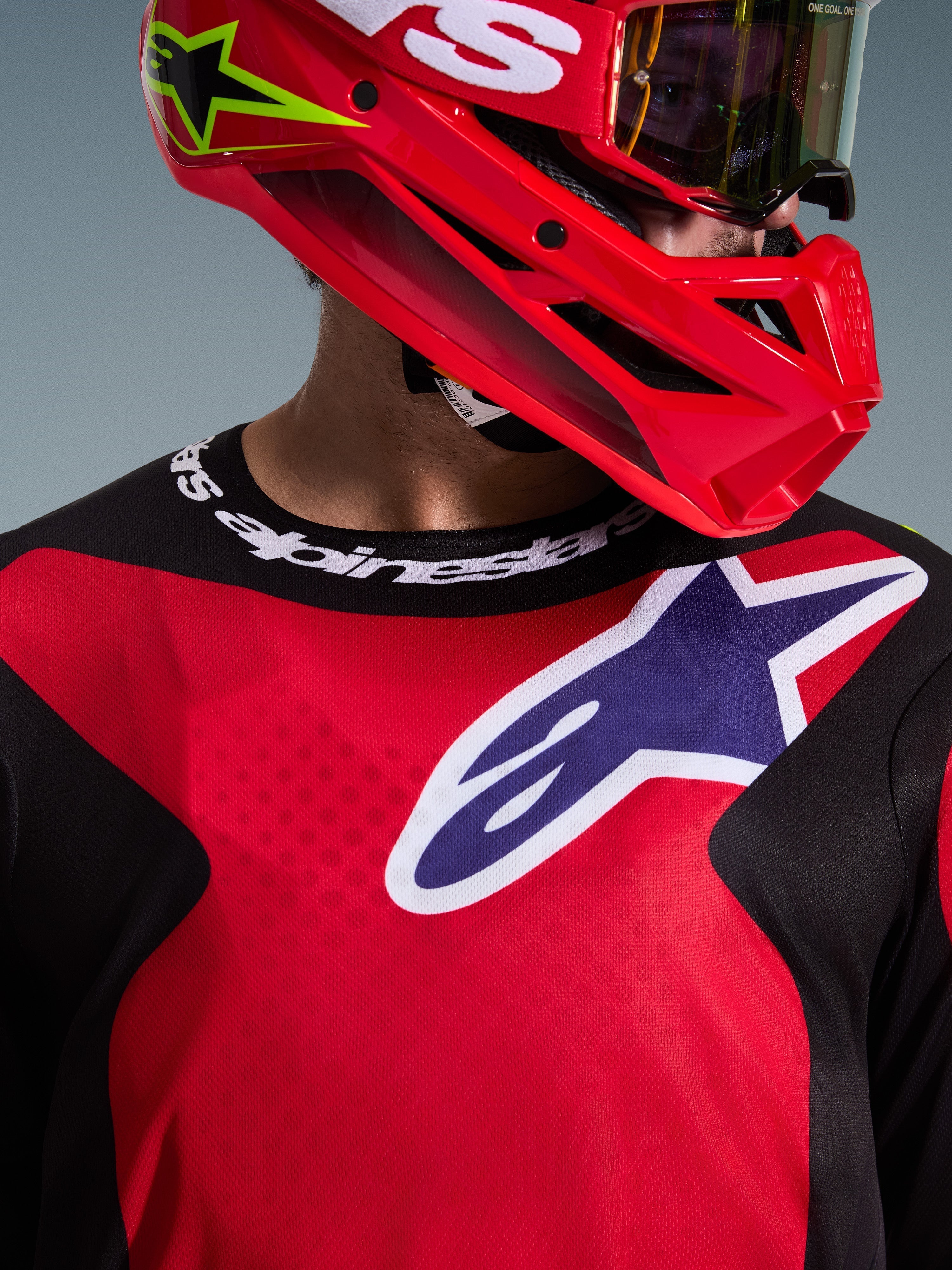 Close-up de um piloto de motocross a usar a Camisola Fluid Grid 2026 em Vermelho, Preto e Roxo, com um logótipo Alpinestars grande no peito e gola desportiva da marca, combinada com um capacete e óculos vermelhos, destacando o polifabric de performance e o ajuste atlético.