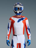 Piloto de motocross com a Camisola Alpinestars Fluid Grid 2026 em Orange Ucla Blue, peito branco com painéis laterais cor de laranja e logótipos azuis, calças brancas e azuis a condizer, luvas azuis e capacete branco e azul, visual profissional de corrida todo-o-terreno