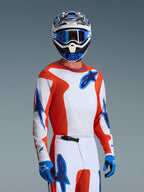 Camisola Alpinestars 2026 Fluid Grid, camisola de motocross em cor de laranja e azul UCLA Blue, usada por um piloto com calças brancas e cor de laranja a condizer, capacete azul e branco, óculos de proteção azuis e luvas azuis, conjunto completo de equipamento de corrida todo-o-terreno