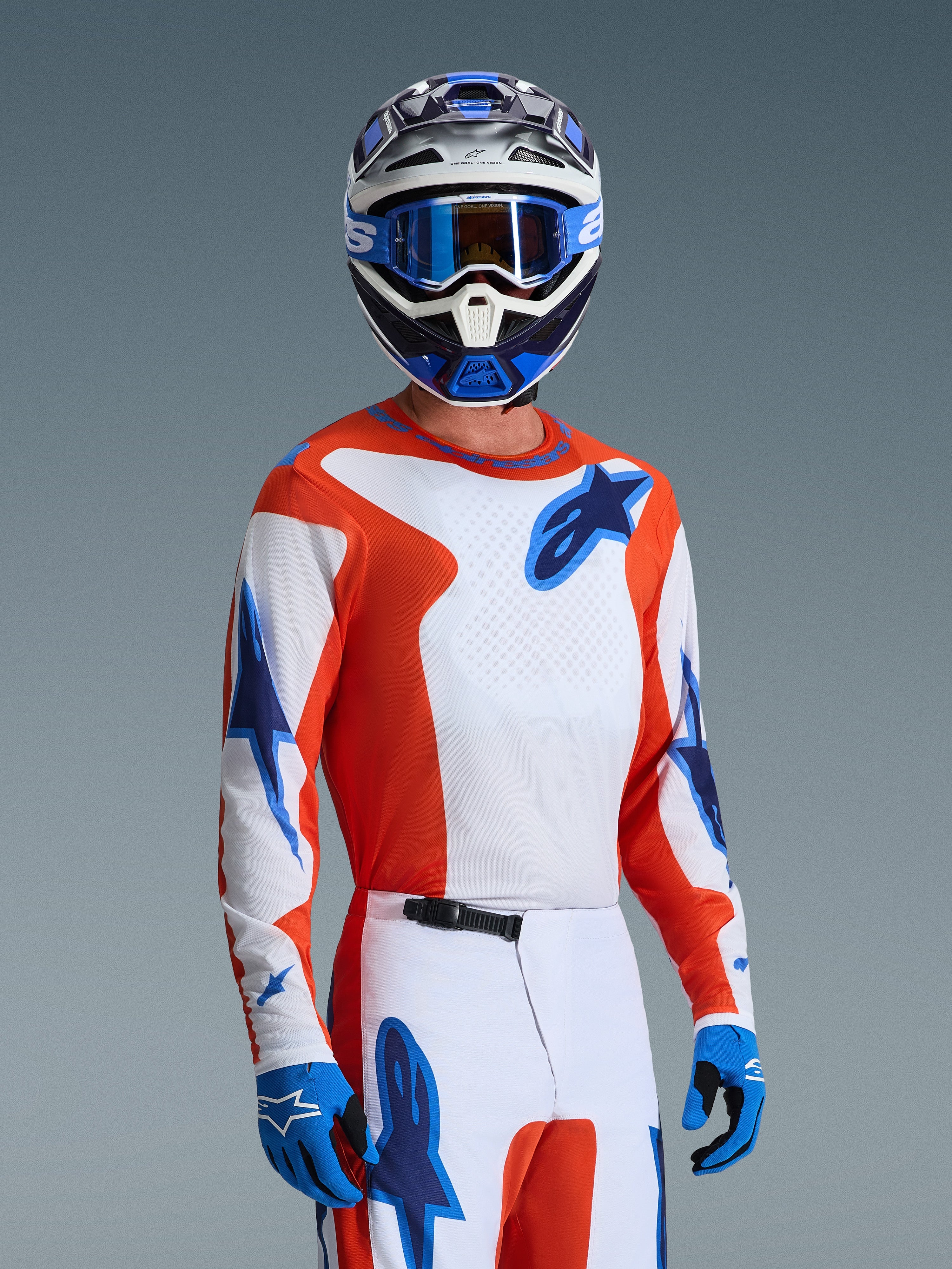 Camisola Alpinestars 2026 Fluid Grid, camisola de motocross em cor de laranja e azul UCLA Blue, usada por um piloto com calças brancas e cor de laranja a condizer, capacete azul e branco, óculos de proteção azuis e luvas azuis, conjunto completo de equipamento de corrida todo-o-terreno