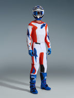 Piloto de motocross com o Conjunto de Equipamento Alpinestars Fluid Grid 2026, camisola e calças brancas com grafismos em laranja e azul UCLA, capacete e óculos azuis a condizer, luvas azuis e botas de corrida azul vivo, conjunto de performance coordenado para condução todo-o-terreno.