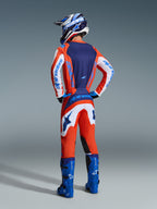 Piloto de motocross visto de costas a usar a camisola e calças 2026 Fluid Grid em laranja e azul UCLA, com mangas brancas com logótipo Alpinestars em azul, botas de motocross em azul brilhante e capacete a combinar, conjunto de equipamento completo para corridas todo-o-terreno.