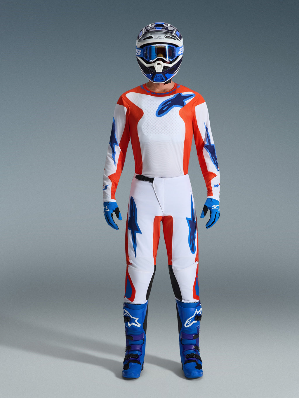 Conjunto de equipamento Alpinestars 2026 Fluid Grid, piloto de motocross com equipamento completo, camisola e calças brancas com detalhes em laranja e azul UCLA, capacete azul e branco a condizer com óculos azuis, luvas e botas de motocross azuis, fato de corrida off-road coordenado