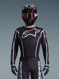 Camisola Alpinestars Fluid Apex 2026, camisola de motocross, preta e cinzenta, com um grande logótipo Astars branco no peito e detalhes gráficos cinzentos nas mangas, usada por um piloto com óculos cor-de-rosa e capacete preto, equipamento de corrida todo-o-terreno coordenado