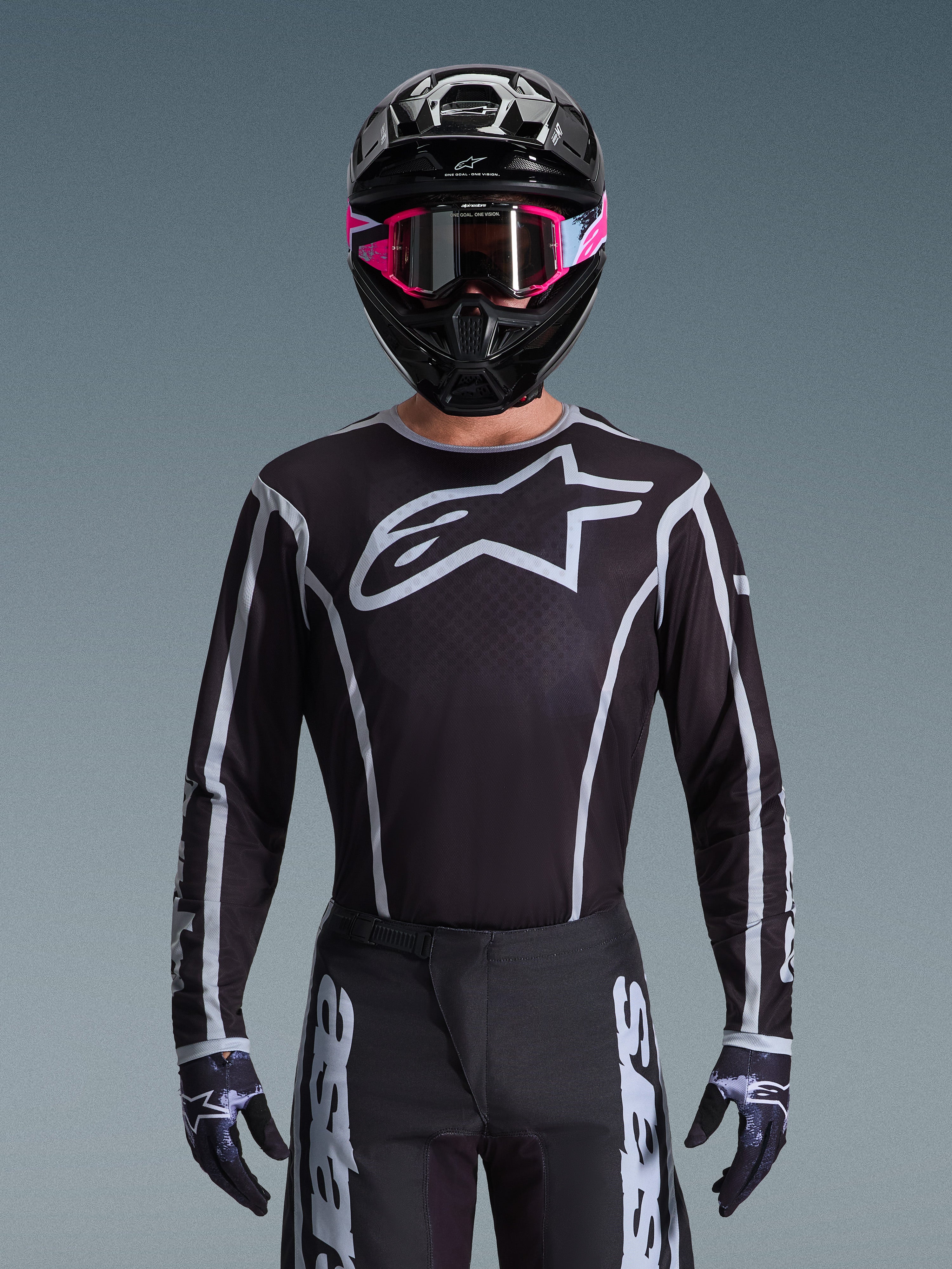 Camisola Alpinestars Fluid Apex 2026, camisola de motocross, preta e cinzenta, com um grande logótipo Astars branco no peito e detalhes gráficos cinzentos nas mangas, usada por um piloto com óculos cor-de-rosa e capacete preto, equipamento de corrida todo-o-terreno coordenado