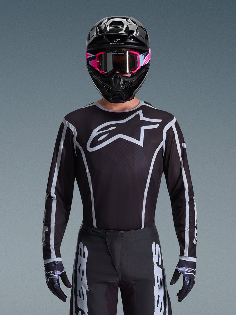 Camisola Alpinestars Fluid Apex 2026, camisola de motocross, preta e cinzenta, com um grande logótipo Astars branco no peito e detalhes gráficos cinzentos nas mangas, usada por um piloto com óculos cor-de-rosa e capacete preto, equipamento de corrida todo-o-terreno coordenado