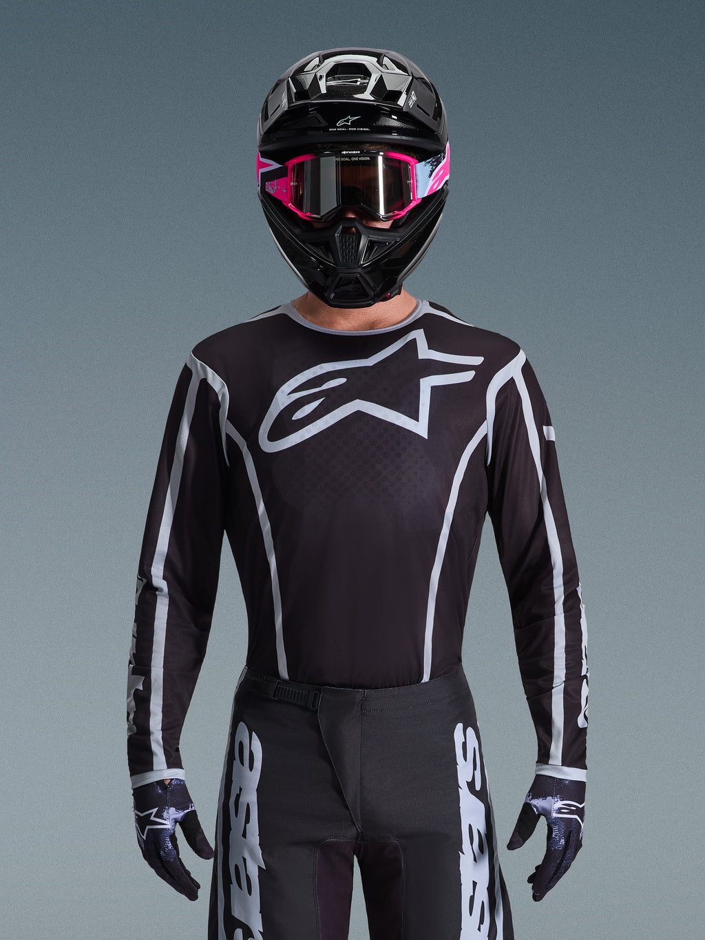 Conjunto de Equipamento Alpinestars 2026 Fluid Apex, camisola e calças de motocross em preto e branco, apresenta um logótipo grande no peito e designs de linhas geométricas, usado com um capacete preto e óculos cor-de-rosa, equipamento de performance para corridas de motocross todo-o-terreno.