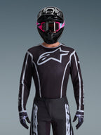 Conjunto de Equipamento Alpinestars 2026 Fluid Apex, camisola e calças de motocross em preto e branco, apresenta um logótipo grande no peito e designs de linhas geométricas, usado com um capacete preto e óculos cor-de-rosa, equipamento de performance para corridas de motocross todo-o-terreno.