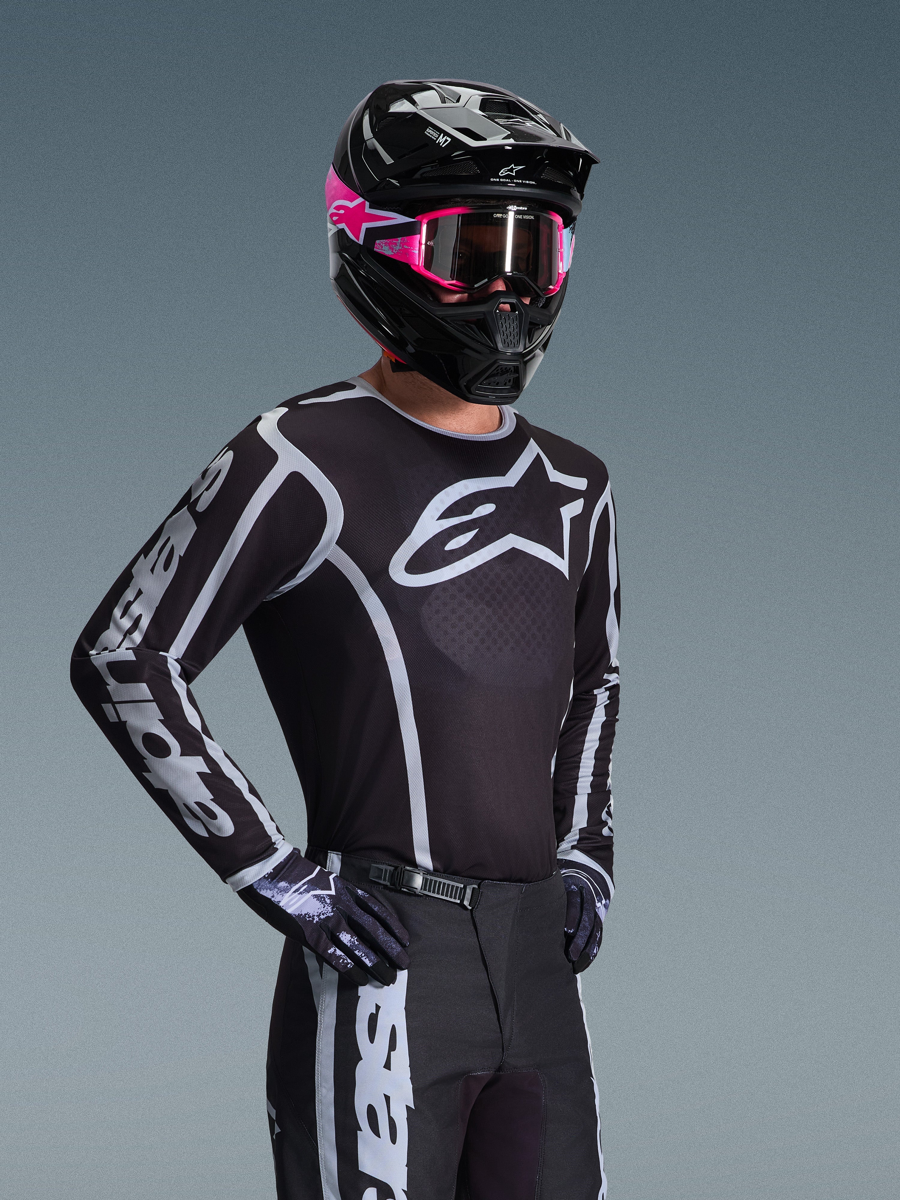 Piloto de motocross a usar a camisola Alpinestars 2026 Fluid Apex, preta e cinzenta com grande logótipo branco no peito e grafismos geométricos nas mangas, acompanhada de calças pretas, luvas pretas e um capacete preto com detalhes e óculos cor-de-rosa