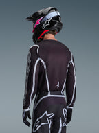 Piloto de motocross visto de costas a usar a Camisola Alpinestars 2026 Fluid Apex em preto e cinzento, calças de motocross a condizer com pormenores brancos, capacete preto com marca em rosa, conjunto coordenado para corridas off-road