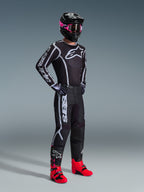 Piloto de motocross a usar a Camisola Fluid Apex 2026, em preto e cinzento, combinada com calças a condizer, capacete preto com óculos cor-de-rosa e botas vermelho vivo, com o logótipo Alpinestars em branco no peito e nas mangas, equipamento de corrida todo-o-terreno profissional coordenado.