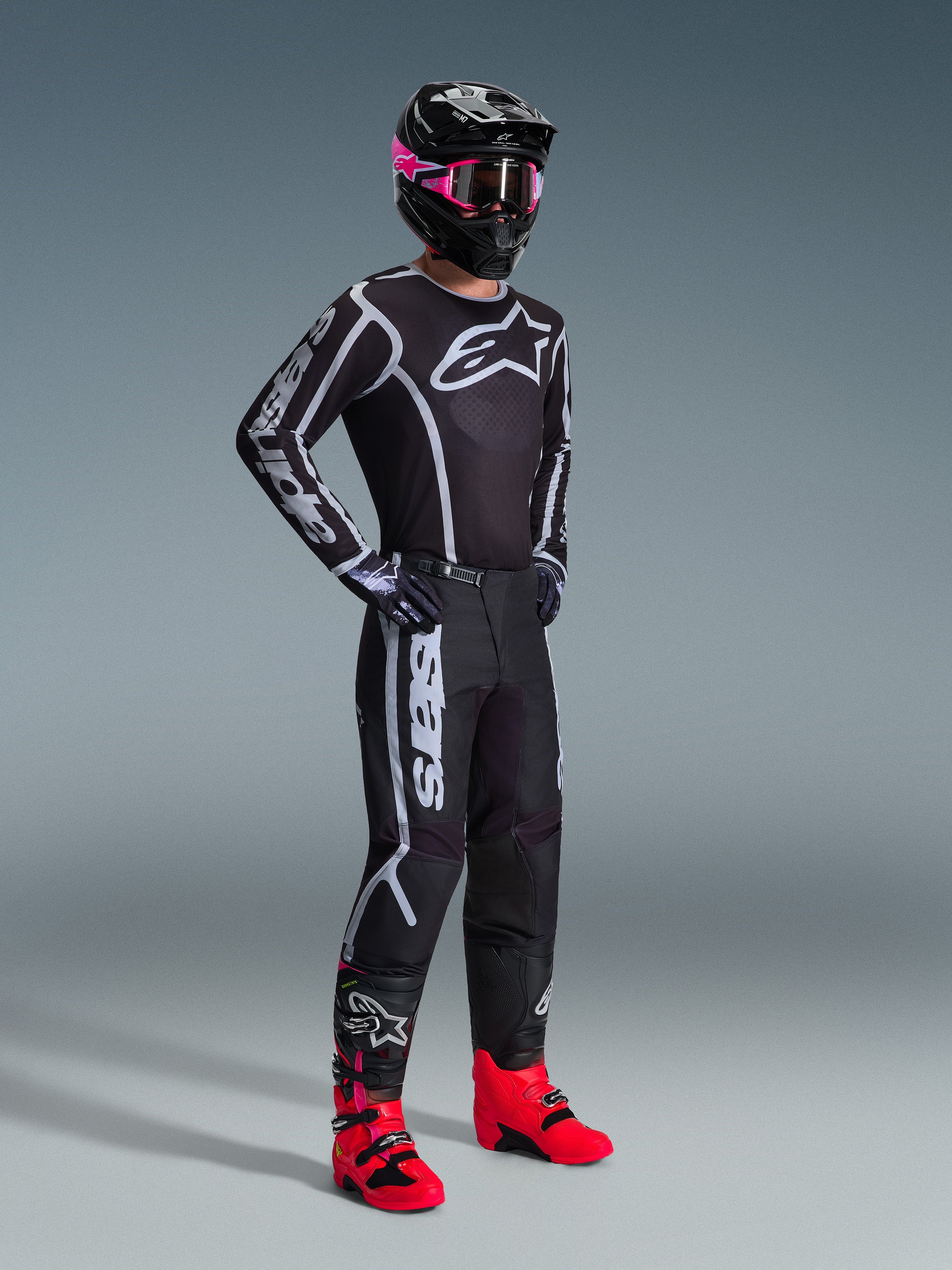 Alpinestars 2026 Fluid Apex Gear Set, piloto de motocross com camisola e calças pretas com detalhes gráficos brancos e cinzentos, capacete preto com óculos cor-de-rosa e botas de corrida cor-de-rosa vibrante, conjunto profissional de motocross coordenado