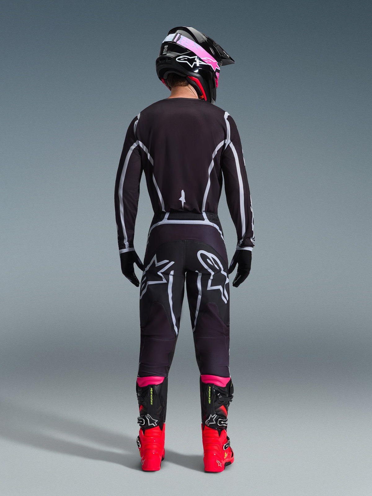 Motocross rider from the back wearing the Alpinestars 2026 Fluid Apex Jersey and matching pants in Black/Gray, featuring white geometric design lines and star logos on the seat, accessorized with a black and pink helmet and red and black racing boots.