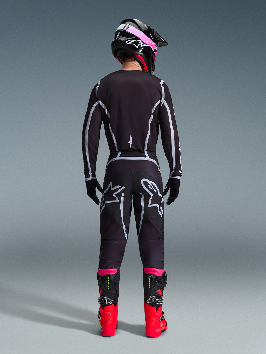 Piloto de motocross visto de costas com a Camisola Alpinestars 2026 Fluid Apex e calças a condizer em preto e cinzento, apresentando linhas geométricas brancas e logótipos de estrela na zona do assento, com capacete preto e cor-de-rosa e botas de motocross vermelhas e pretas.