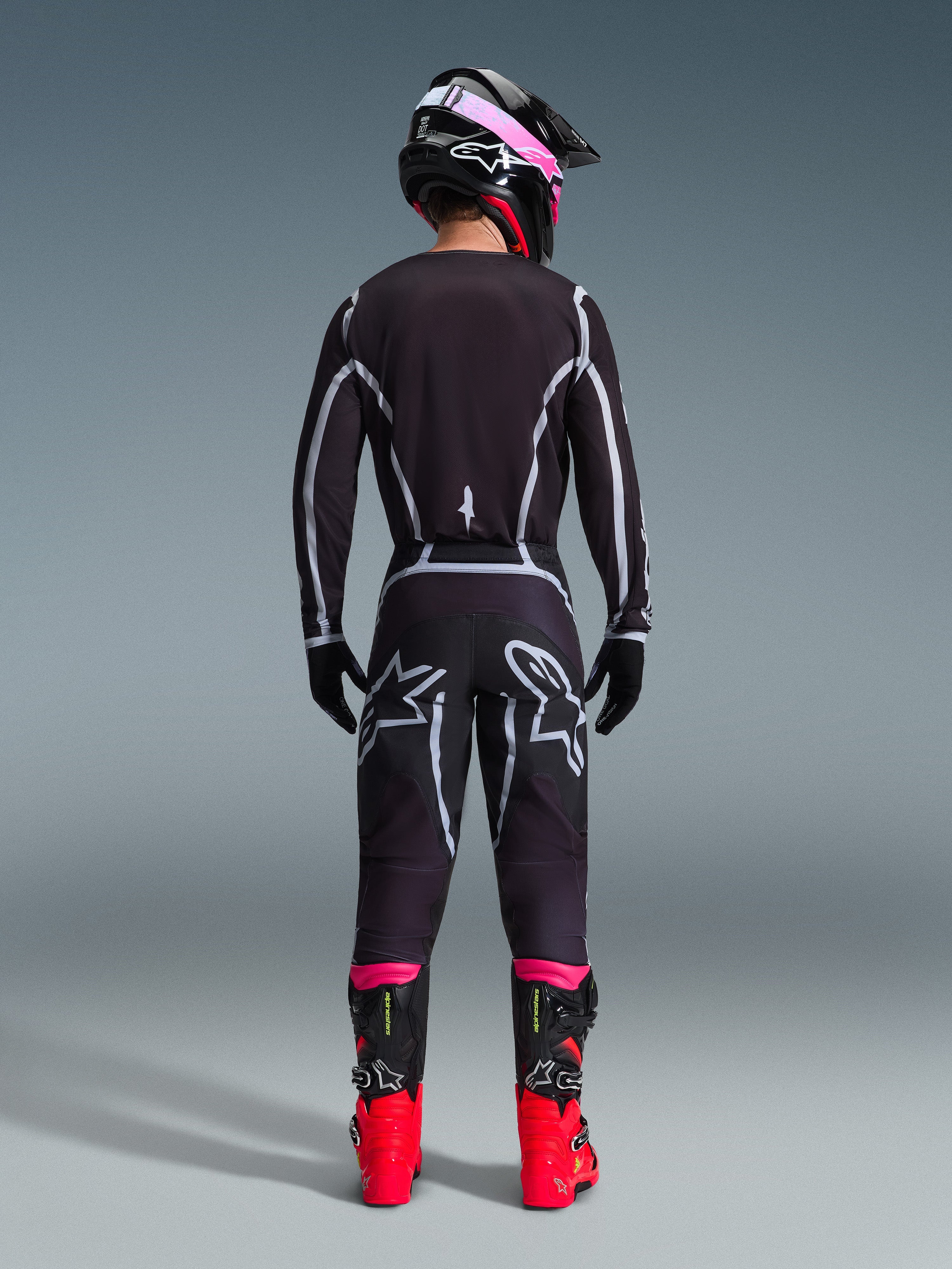 Piloto de motocross visto de costas com a Camisola Alpinestars 2026 Fluid Apex e calças a condizer em preto e cinzento, apresentando linhas geométricas brancas e logótipos de estrela na zona do assento, com capacete preto e cor-de-rosa e botas de motocross vermelhas e pretas.