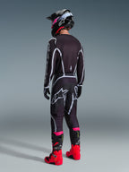 Piloto de motocross a usar o Conjunto de Equipamento Alpinestars Fluid Apex 2026, camisola e calças pretas com linhas geométricas brancas e cinzentas, vista traseira, combinado com um capacete preto com detalhes cor-de-rosa e botas de corrida vermelhas e cor-de-rosa vivas, fotografia de estúdio sobre um fundo cinzento.