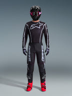 Piloto de motocross a usar a camisola Alpinestars 2026 Fluid Apex, preta e cinzenta, com grande logótipo Astars branco no peito, linhas geométricas brancas, calças pretas a condizer, capacete preto com óculos cor-de-rosa e botas cor-de-rosa vibrante, equipamento completo de motocross