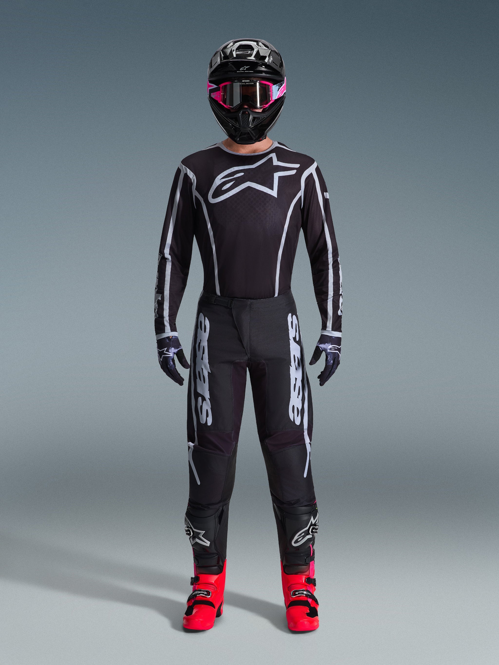 Motocross rider wearing the Alpinestars 2026 Fluid Apex Jersey, Black and Gray, large white Astars logo on the chest, white geometric lines, matching black pants, black helmet with pink goggles, and bright pink boots, full motocross racing outfit
